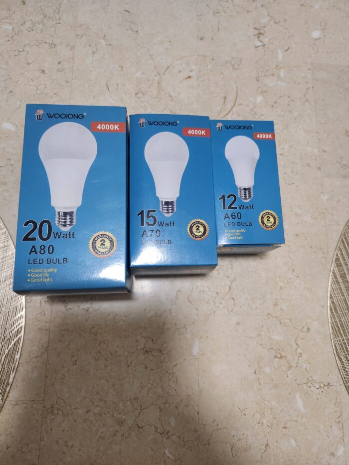 Three blue WooJong LED bulb boxes arranged on a beige tiled floor: 20W A80, 15W A70, and 12W A60, all with bulb illustrations and 4000K. - Tradedubai.ae Wholesale B2B Market