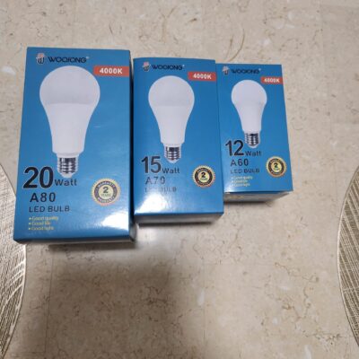 Three blue WooJong LED bulb boxes arranged on a beige tiled floor: 20W A80, 15W A70, and 12W A60, all with bulb illustrations and 4000K. - Tradedubai.ae Wholesale B2B Market