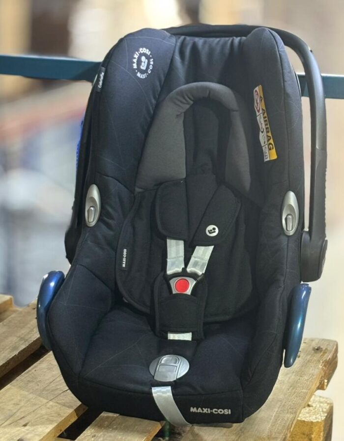 Black Maxi-Cosi infant car seat with five-point harness and buckle, placed on a wooden pallet. - Tradedubai.ae Wholesale B2B Market