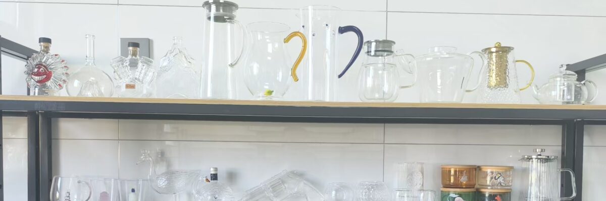 Assorted clear glassware—teapots, pitchers, and jars—on a metal shelf with a few colored-handle pieces (yellow and purple).
